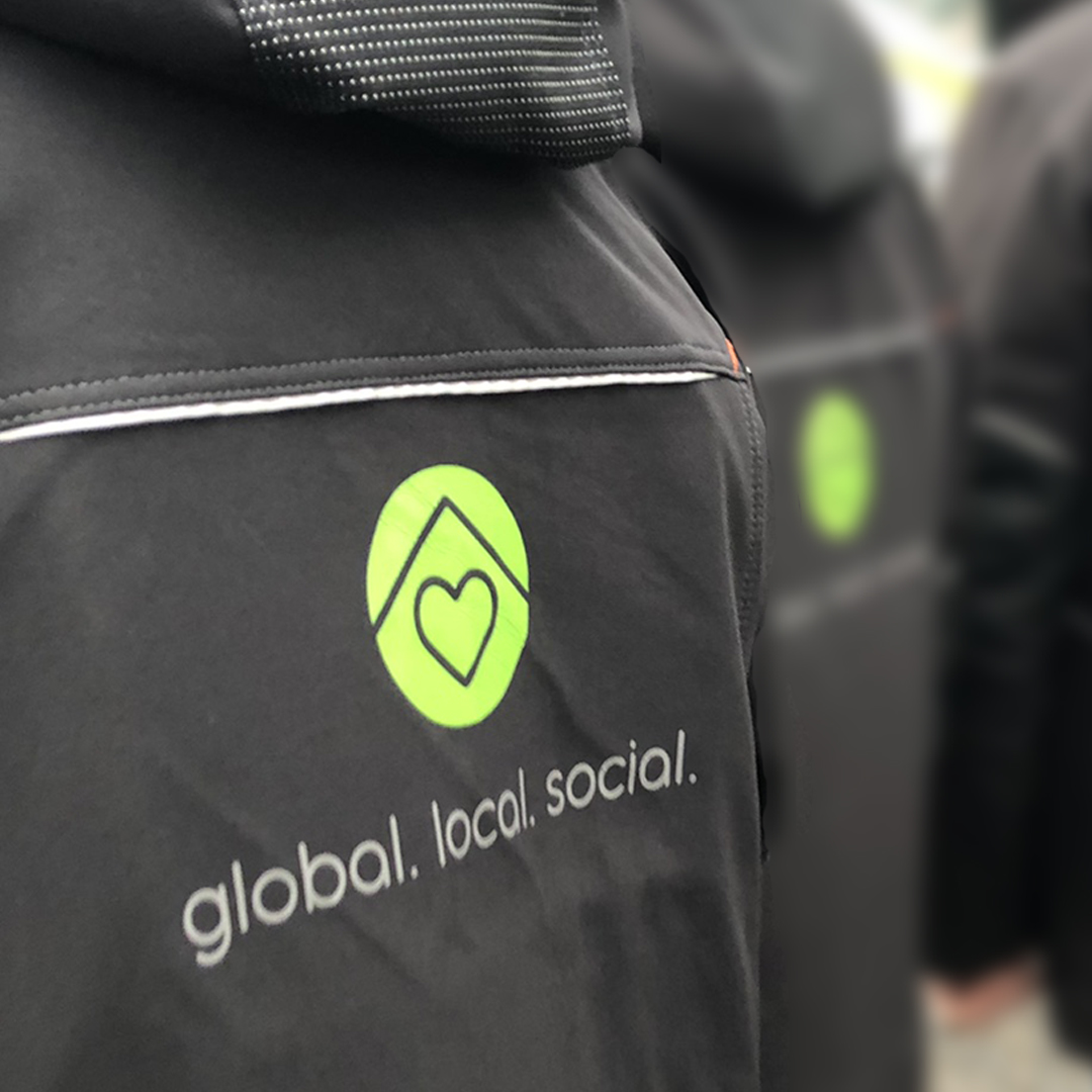 Eine Rückseite von einer schwarzen Arbeitsjacke, wo ein kleine Reflexpaspel eingebaut ist und auf dem Rücken ist das Logo global.local.social aufgedruckt